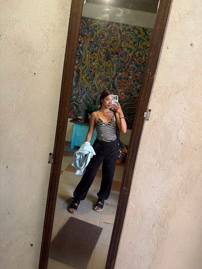 A woman in a black and white zebra-print tank top and black pants is taking a mirror selfie. She is holding a light blue garment in her left hand and a black bag is slung over her right shoulder. She is wearing black sandals. The mirror reflects a colorful mural on the wall behind her, as well as a turquoise cabinet and a brown mat on the tiled floor. The scene appears to be indoors, likely in a residence, as indicated by the wall texture and tiling. Tejakula, Indonesia is a coastal village on the northern side of Bali, known for its tranquil beaches and traditional Balinese culture.