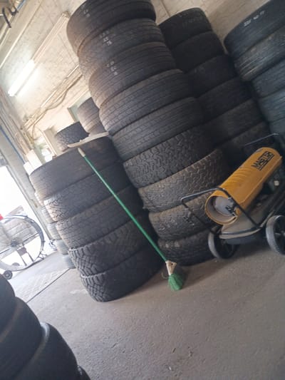 This indoor scene shows stacks of tires and a portable heater, likely in a storage area or workshop in Clarksville, Tennessee, USA. Numerous tires are stacked, some bearing visible markings. A yellow "MASTER" branded portable heater on wheels is positioned on top of some tires. A green-handled broom leans against the tire stacks. The surroundings include a ceiling with fluorescent lighting.