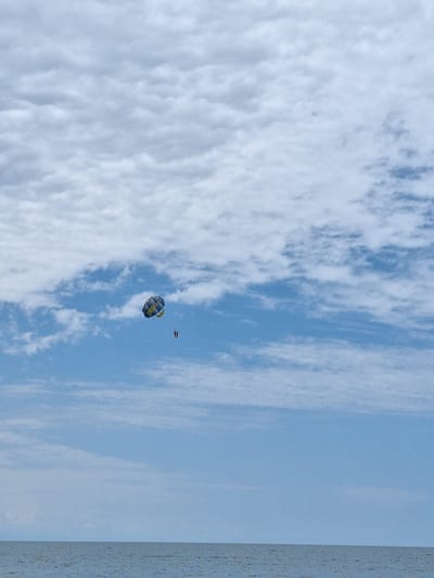 The image depicts a bright, sunny day with a clear blue sky partially obscured by wispy white clouds. The scene is dominated by the vast expanse of the sky, with a hint of the sea on the right side of the frame, indicated by a textured, light blue line. In the middle ground, a person is parasailing, suspended in the air by a colorful parachute. The parachute displays a pattern of blue, yellow, and white sections. Directly behind the parachute, a smaller dark object, likely a boat towing the parasailor, is visible. The overall mood is one of leisure and outdoor activity, suggesting a recreational setting likely at a beach or coastal resort. The time of day appears to be midday, given the bright lighting and clear sky. No text is visible in the image.