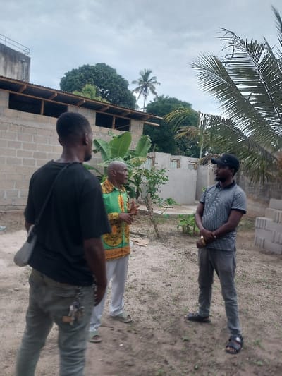 This image depicts three adult males engaged in a conversation in an outdoor, unpaved area in Mwandege, Tanzania, under an overcast sky. The foreground shows dirt ground with sparse low-lying vegetation. To the left, a male subject with his back to the viewer wears a black t-shirt, light-colored jeans with keys clipped to the right hip, and a light grey shoulder bag. Central to the frame, an older male, facing left, wears a green and gold patterned long-sleeved shirt, light trousers, and light-colored open-toed sandals; he is actively gesturing with both hands. To the right, a third male, facing the center, wears a dark baseball cap, a grey and black striped polo shirt, dark trousers, and black open-toed sandals; his hands are clasped at waist level, appearing to hold a gold-colored object. In the background, a building constructed of concrete blocks with a corrugated metal roof and exposed wooden rafters is visible. Behind this structure, dense green foliage, including a banana plant and palm trees, is present. Additional concrete blocks are stacked on the far right. A water tank sits atop a distant building on the left.