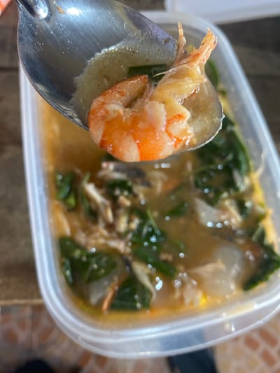 The image shows a close-up view of a spoon lifting a cooked shrimp from a clear plastic container filled with a light-colored soup. The soup contains visible pieces of green leafy vegetables, possibly spinach or kangkung, and shredded meat, which could be chicken or fish. The shrimp is orange and appears to be cooked through. The container is placed on a surface that appears to be a wooden table, with the background slightly out of focus. The setting suggests a casual meal, likely in a residential or food stall environment within Ta', Indonesia, though no specific landmark can be identified to pinpoint the exact location.