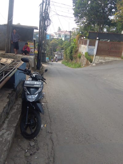 The image depicts a street scene in Cisarua, Indonesia, likely in a residential or mixed-use area. A black Honda motorcycle with license plate F 3098 FDK is parked on the side of a paved road. The motorcycle has a black frame, black wheels, and a black seat. The license plate is white with black lettering and numbers. A cigarette butt is visible on the ground near the motorcycle's front wheel.
To the left of the road, there is a raised concrete area with a small, open-fronted structure, possibly a shop or shelter, with a corrugated metal roof. Two individuals are present in this area. One, a person wearing a plaid shirt and dark shorts, is standing and leaning against the structure. Another person, wearing a red jacket and dark pants, is sitting on the edge of the concrete. A yellow sign with red text that reads "JUAL POTON" is visible behind the sitting person, suggesting a sale of something related to "poton".
Further down the road, several other motorcycles are seen moving in the distance. The road curves to the right and ascends a hill. On the right side of the road, there is a stone wall supporting a higher ground area with lush green vegetation. A gate with ornate metalwork and a dark fence are visible at the top of the slope. Buildings, including a multi-story structure with white walls and a blue roof, are visible in the background, suggesting a town or village setting. The overall environment appears to be an outdoor, daytime scene with moderate sunlight.