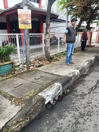 A man in a cap, vest, and jeans stands on a sidewalk next to a tree, looking towards another man in a maroon jacket. A restaurant sign is on a pole behind the tree. A white fence borders a building with a red roof. A gutter with visible damage runs along the edge of the sidewalk. Sewer covers with metal handles are visible on the sidewalk. Parked motorcycles can be seen behind the fence.