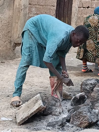 An outdoor scene in Rangaza, Nigeria shows a young man bent over, focused on an activity on the ground. He wears a light blue, long-sleeved tunic and matching trousers, with light-colored slide sandals on his feet. His hands are near a torn, reddish-brown bag, suggesting he is either filling it, emptying it, or manipulating its contents, possibly within or around a pile of dark rocks and ash in the foreground.
To the right, partially visible, stands another person wearing a patterned skirt with circular motifs, colorful striped socks (orange, green, pink, blue), and dark sandals. The ground is dusty or sandy. In the background, a weathered building or wall with exposed brick or mud-like textures can be seen. The lighting suggests it is daytime. The man's expression is one of concentration as he looks down at his hands and the task before him. There is no discernible text visible in the image.