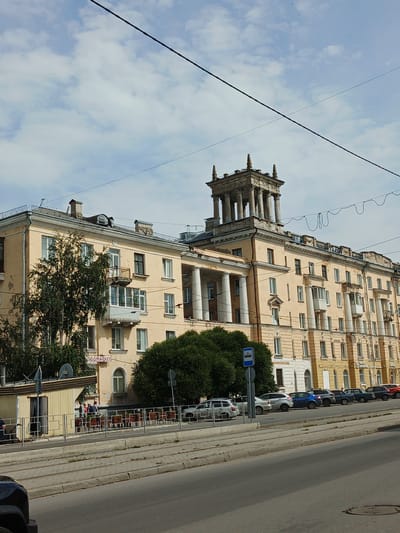 The image captures a street scene in Zlatoust, Russia, focusing on a large, pale yellow, multi-story apartment building with classical architectural elements, including prominent white columns. The building exhibits a somewhat weathered facade. A distinctive tower-like structure with columns and decorative elements rises from the rooftop on the right side. In front of the building, a row of parked cars is visible along the sidewalk. A small kiosk or shop with a yellow facade is situated to the left, near a tree. People are gathered near the kiosk and along the fence in front of the building. A bus stop sign is also present. The street in the foreground shows tram tracks, indicating a public transportation route. Overhead, power lines crisscross the sky, which is partly cloudy.