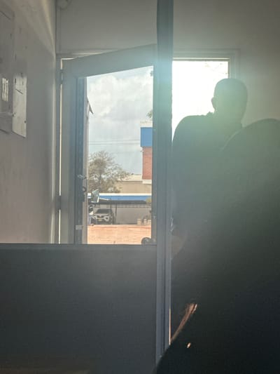 The image captures a view through an open doorway, looking outwards into an urban environment. The foreground is dominated by a dark, out-of-focus mass, possibly a person or furniture, obscuring the lower right portion of the scene. The doorway itself is framed by a light-colored wall on the left and a bright, vertical metal beam on the right. Sunlight streams in from the right, creating a strong glare and silhouetting a person's head and shoulders visible in the middle ground, standing just beyond the doorway. This person appears to be wearing dark clothing.
Through the doorway, the exterior scene shows a paved area with a white SUV parked under a covered structure. Beyond the car, there is a low building with a white wall and a blue trim. Further in the background, a bare-branched tree is visible against a bright, cloudy sky. The overall impression is a moment captured in daylight, with significant contrast between the brightly lit exterior and the darker interior. The scene is set in the city of Juan Mina, Colombia.