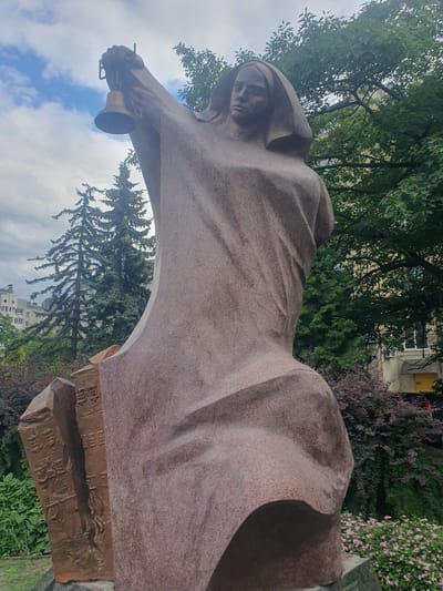 The image shows a full shot of a statue in Ivano-Frankivsk, Ukraine. The statue is made of pink granite and depicts a woman wearing a shroud and holding a bell in her right hand, raised above her head. The woman's head is bowed and her expression is solemn. The statue is placed in a garden setting with trees, bushes, and small pink flowers. In the background, there are apartment buildings and a cloudy sky. The statue is situated in front of a large, rectangular stone with embossed text, possibly a memorial plaque. The overall impression is one of solemnity and remembrance.