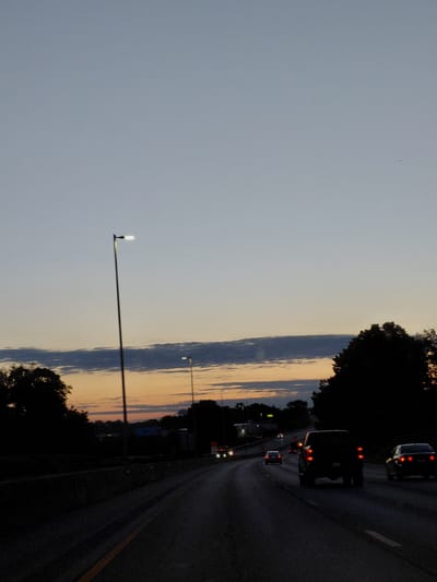 This is a vertically oriented, eye-level, medium shot photograph taken at dusk on a highway in Lexington, United States. The sky is a gradient of light blue fading into warmer tones of orange and pink near the horizon, suggesting the sun has recently set. A few dark, elongated clouds streak across the sky. The foreground is dominated by the dark asphalt of the highway, with a yellow line marking the edge of the travel lane. A few cars are visible on the highway, their brake lights glowing red, indicating slow-moving traffic or a stop. One car in the foreground has its headlights on.

To the left of the highway, there is a tall street lamp with a bright, white light. Further to the left, the sky takes up a significant portion of the frame, with a tiny, dark object that could be a bird or insect visible in the upper left quadrant. Silhouetted trees line the right side of the highway, their dark shapes creating a stark contrast against the colorful sky. The overall mood is calm and serene, with a touch of melancholy associated with the end of the day. There is no visible text in the image.