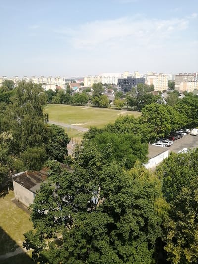 The image shows a panoramic view of a residential area in Salihorsk, Belarus. In the foreground, a large building with a grey roof is visible, surrounded by lush green trees. Beyond the building, a sports field with a running track and various sports equipment can be seen. Further in the background, a row of multi-story apartment buildings stretches across the horizon, characteristic of Soviet-era architecture. A parking lot filled with several cars and a white van is situated near some of the apartment buildings. The sky is clear and blue, with a few scattered clouds, indicating a sunny day. The overall scene depicts a typical urban landscape with residential blocks, green spaces, and recreational facilities.