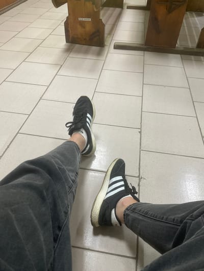 The image depicts a close-up view of a person's legs and feet, clad in dark grey jeans and black Adidas sneakers with white stripes. The person is seated, with their legs crossed, on a floor tiled with light-colored, square tiles. In the background, wooden benches are visible, suggesting a public space such as a waiting area, a church, or a lecture hall. A label with the text "Mod. No. 189201140" is affixed to one of the wooden benches. The lighting is neutral and the scene is unadorned, focusing solely on the footwear and lower limbs of the individual. There is no specific indication of the location being in the city of Varash, Ukraine, beyond the prompt's request to include this information.