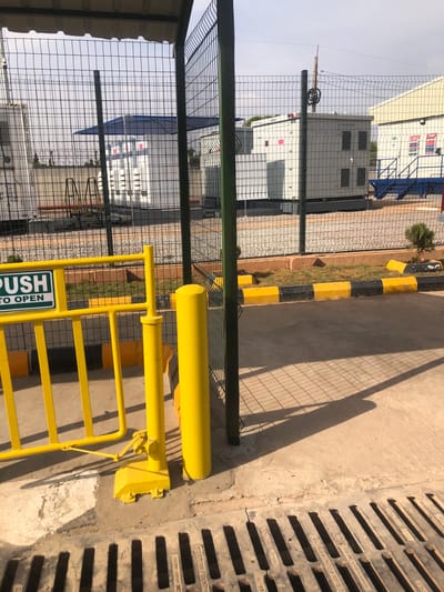 The scene depicts an outdoor industrial facility entrance in Nassarawa, Nigeria. In the immediate foreground, a concrete surface features a metal grate drain with visible inscriptions "STEEL FOUNDRIES" and "GERMAN FOUNDRIES". Strong shadows from fencing and other structures are cast across the concrete.
A bright yellow pedestrian barrier, partially open, is positioned centrally in the left foreground. Attached to this barrier is a green sign with white text reading "PUSH TO OPEN". Adjacent to the barrier, a cylindrical yellow bollard stands upright.
Beyond the foreground elements, a dark green metal mesh fence forms a primary barrier. Behind this fence, a landscaped strip features brown soil, sparse dry grass, and small green shrubs, bordered by a curb painted in alternating yellow and black sections.
Through the fence, several white modular structures or industrial units are visible on a gravel ground. These include a large white unit with ventilation grates, likely a generator or machinery enclosure. Another white building-like unit has a blue roof or tarp covering part of its top. A blue metal staircase is visible leading up to one of these structures. Barbed wire can be seen atop a perimeter wall in the distance. The sky above is light blue with some haziness. No individuals are present in the image.