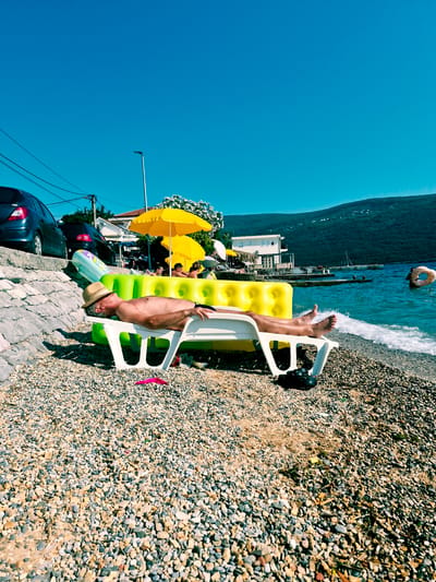 A man wearing a straw hat lies on a white plastic beach lounger on a pebble beach. He is shirtless and appears to be sunbathing. Behind him, a bright yellow inflatable structure is partially visible, with an inflatable mattress resting on top. Further back, two yellow beach umbrellas are set up. In the background, there are cars parked, buildings, trees, and a hillside covered in greenery, characteristic of the coastal scenery in Kumbor, Montenegro. The Adriatic Sea is calm and blue, with small waves lapping at the shore. The sky is clear and bright blue.