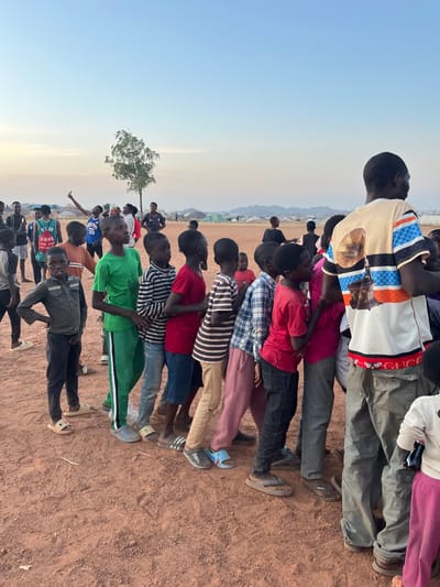 A horizontal outdoor shot displays a group of children lined up in rows. Most have dark skin and short hair. They wear varied casual clothing, like T-shirts, pants, and shoes. A few adults stand behind the children. The ground is reddish-brown soil, and a lone tree is nearby. The sky is a gradient of light blue. The children are looking up, likely at a person or event just out of view.