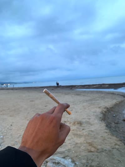 The image depicts a close-up of a right hand, likely male, holding a rolled cigarette or joint against a coastal backdrop in M'diq, Morocco. The hand, with visible veins, holds the light-colored, rolled item which has a distinct white filter tip.

The scene is set on a sandy beach, stretching along a calm body of water, likely the sea. The horizon line is visible where the water meets a soft, overcast sky, filled with light blue-grey clouds. This suggests either early morning or late afternoon/evening due to the diffused lighting conditions.

In the far distance, near the water's edge on the beach, two small, indistinct human figures can be observed, adding a sense of scale and presence to the vast landscape. The overall mood conveyed by the muted colors and cloudy sky is tranquil. There is no visible text in the image.