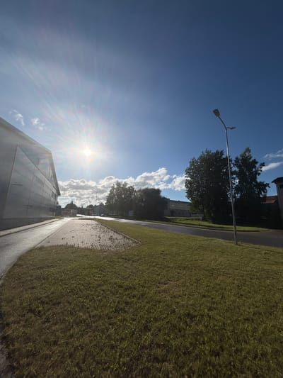 The image depicts a street scene in Sauleskalns, Latvia, under a bright, sunny sky with scattered clouds. The foreground features a well-maintained lawn bordering a paved walkway and a road. A modern building, possibly an industrial or commercial facility, occupies the left side of the frame, its metallic facade reflecting the sunlight. In the middle ground, a solitary lamppost stands near a cluster of mature trees. The background reveals more buildings and foliage, suggesting a developed urban or suburban area. The sun is intensely bright, casting lens flares and creating a radiant effect across the sky, indicating it is likely midday. No people are visible in the scene.