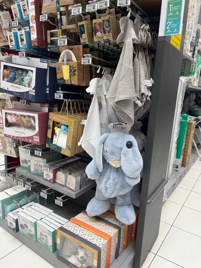 The image shows a section of a retail store in Saint-Brice-Courcelles, France, located at latitude: 49.283, longitude: 3.999. The store aisle features shelves stocked with various baby products. On the left side, there are several boxes of baby toys, including musical mobiles and plush toys. On the right side, there is a large display of baby blankets and swaddles. In the center, a large, blue plush bunny with a button eye sits on a lower shelf, partially obscured by a grey blanket. Below the bunny, there are several boxes of baby clothes, including a box labeled "PLAID DOUDOU" and a box with a picture of a baby wearing a white onesie. The overall scene depicts a typical retail environment showcasing baby products.