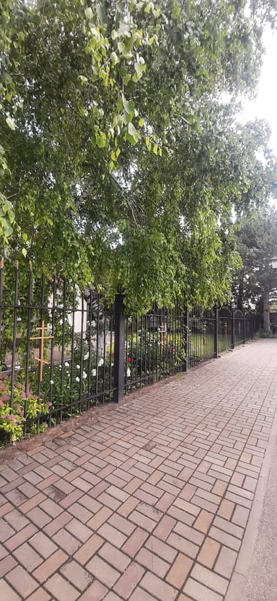 The image depicts a paved walkway with a brick pattern, bordering a black metal fence. Beyond the fence, there is a garden with flowers and trees. Two wooden crosses are visible attached to the fence. The scene is likely in a cemetery or memorial park, given the presence of crosses and flowers. The overall setting is outdoors, with lush greenery and a cloudy sky. The image's context suggests it is located in Yessentukskaya, Russia, potentially a place of remembrance.