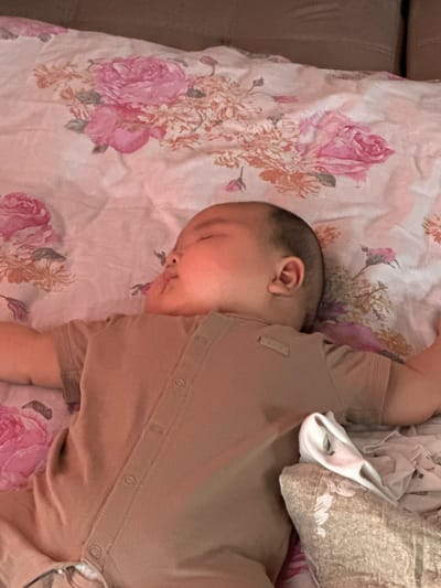 A baby is sleeping on a bed with a floral patterned sheet. The baby is wearing a tan colored onesie. The sheet has pink roses and other flowers. A blanket is visible behind the baby. The scene appears to be indoors. There is no visible text.