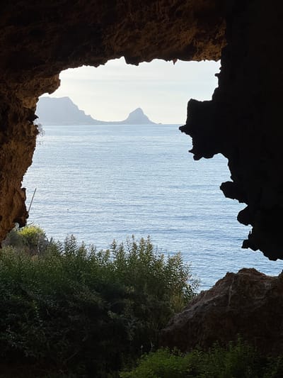 The image presents a view from within a dark rock formation, likely a cave or overhang, overlooking the sea and distant land in Sperone, Italy. The immediate foreground consists of jagged, dark brown rock formations, textured with visible strata and irregularities, framing the scene. Below the rock opening, dense, dark green foliage, including tall, leafy plants and shrubs, partially obscures the immediate terrain. A thin, light-colored pole extends vertically from the vegetation on the lower left.
The midground is dominated by a calm, blue-grey body of water with subtle ripples on its surface. A small, spherical red object, possibly a buoy, floats in the water slightly to the left of the central axis. In the background, across the water, distant landmasses composed of hazy, desaturated mountains or large hills are visible under a light grey-blue sky, which appears brighter towards the horizon. One prominent, conical peak is visible on the right side of the distant land. No individuals, animals, or additional man-made structures are discernible within the frame.