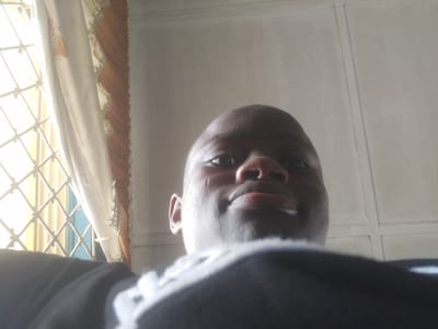 A low-angle close-up photograph depicts a person's face, likely a young adult, with dark skin, focused on their eyes, nose, and mouth. The individual's head is mostly bald. The subject wears a dark-colored garment with a white design at the top. Behind the subject, the image reveals a white, textured ceiling with thin, recessed lines. To the left, a window with a white, lattice-like frame is partially covered by a light-colored curtain featuring an orange and beige patterned trim. Given the prompt includes the location of Epe, Nigeria, this scene likely takes place indoors at a residential location within the city.