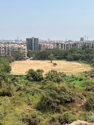 The image shows an open, dirt field in Ambernath, India, bordered by a mix of trees and dense vegetation on one side, and a line of multi-story residential or commercial buildings on the other. Construction appears to be ongoing on some of the buildings. Power lines stretch across the upper left portion of the frame. A red tractor is visible in the middle of the field. A single individual is standing near the tractor. Other yellow construction vehicles are parked by the buildings, next to the field.