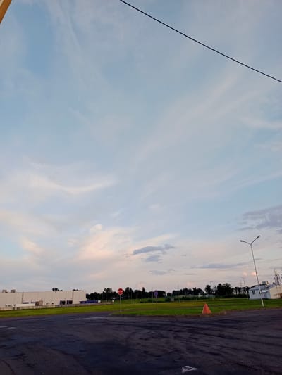 The image depicts an outdoor scene, likely in an industrial or commercial area, under a sky filled with wispy clouds. The foreground is an asphalt surface with tire marks, suggesting it might be a parking lot or a driving area. In the mid-ground, there is a well-maintained green lawn with scattered trees and a few structures. One prominent structure is a large, light-colored industrial building with multiple loading bays. To the right, there is a smaller white building with a light pole in front of it. Various other light poles are visible across the scene. A red stop sign is placed on a pole in the grassy area. An orange traffic cone is also present on the lawn. In the background, more trees and some industrial structures, including what appears to be a power transmission tower, are visible against the horizon. The sky transitions from a light blue at the top to softer hues of pink and white near the horizon, indicating it may be dusk or dawn. A dark power line stretches diagonally across the upper portion of the image. The location is identified as Shchomyslitsky Sel'sky Soviet, Belarus. There are no people visible in the image.