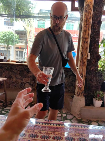 A medium shot captures a man holding a glass of water, within what appears to be a restaurant or cafe setting in Thành phố Hà Giang, Vietnam.
The man has fair skin, a bald head, a beard, and wears glasses. He is dressed in a gray t-shirt and dark shorts, with a black bag slung across his shoulder. He extends the glass of water forward, towards another person whose hand is partially visible in the foreground.
The background features a stone wall, a glass window overlooking a street scene, and a large tree trunk. The street outside the window shows buildings, greenery, and a glimpse of signs in Vietnamese. The interior has a patterned tablecloth, and flooring with a mix of tiles and stones. A small plant in a white pot is also visible.