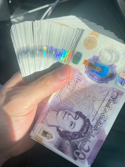 A human left hand holds a fanned stack of new polymer twenty-pound sterling banknotes (GBP). The hand's thumb and index finger are visible, securing the bundle. The top banknote displays the portrait of Queen Elizabeth II on the obverse, with "Twenty Pounds" and "Bank of England" text clearly legible. A prominent vertical holographic strip on the left side of the stack exhibits a rainbow spectrum of colors. A large transparent window on the visible banknote features a metallic foil patch depicting the north-west tower of Lichfield Cathedral, located in Lichfield, United Kingdom. Additional holographic elements within the window include a crown and text alternating between "£20" and "Twenty". The background is out of focus, suggesting an interior environment, possibly a vehicle, with a dark surface beneath the banknotes. Strong, direct lighting illuminates the scene, casting highlights on the banknotes and hand.