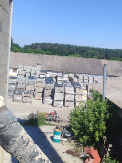 In Bilohorodka, Ukraine, an outdoor industrial area features a large quantity of stacked pavers, or paving stones, of various sizes and colors. The pavers are neatly arranged on wooden pallets, some wrapped in plastic. Behind the stacks of pavers, a long, gray, corrugated roof structure extends, with a white building visible beneath part of it. A metal chimney pipe stands vertically on the right side of the roof. A bright red motorcycle is parked on a patch of grass near the stacks of pavers. In the foreground, a large, black, flexible pipe, possibly insulation or a conduit, is partially visible, draped and covered in dust. A teal barrel with white lettering sits on the ground to the right of the pipe. Beyond the buildings, a dense line of green trees forms the horizon under a clear blue sky.
