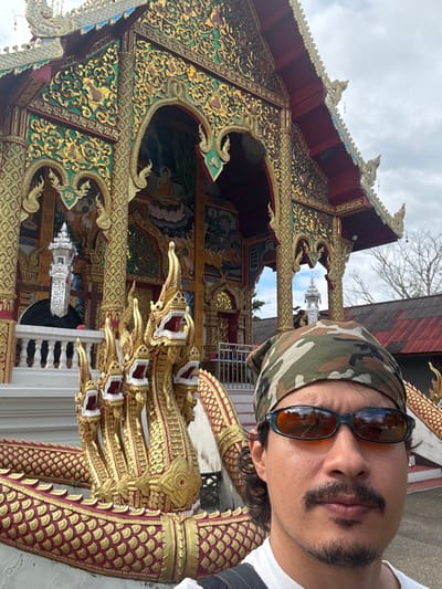 The image displays an outdoor, daytime scene in Ban Mae Na Chalong, Thailand. A male subject occupies the foreground, positioned on the right side of the frame, taking a selfie. He has dark hair, a mustache, and a short goatee, and wears a camouflage-patterned bandana, dark-framed sunglasses with orange-tinted lenses, and a white t-shirt. His expression is neutral.

Behind the subject, a prominent, highly ornate Thai temple structure dominates the background. A large golden, multi-headed Naga statue with red scales and white detailing forms a significant part of the temple's foreground architecture on the left. The temple itself features a steep red roofline and a richly decorated facade, predominantly gold and green with intricate patterns, carvings, and mirrored elements. Multiple arched openings or recesses within the facade reveal colorful murals, including depictions of a seated Buddha on a lotus throne. A distant silver-colored spire or stupa is visible through one of the arches on the right side of the temple. The sky above is overcast. No interactions between individuals are visible.