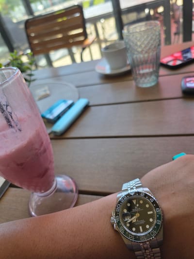 The image shows a person wearing a Rolex watch with a black dial and a jubilee bracelet. Next to the person's arm is a glass of pink smoothie. In the background are a glass of water, sunglasses, a coaster, and a chair. The setting appears to be an outdoor cafe or restaurant. The location is not determinable from the image.