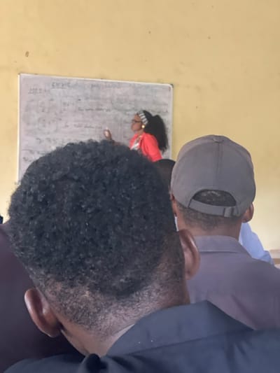 The image depicts an instructional setting in Uturu, Nigeria. A female instructor is prominently featured in the upper mid-ground, standing to the right of a whiteboard and actively writing on it with a white marker. She is wearing a red top, eyeglasses, and a black and white striped headband, with dark, curly hair pulled back. The whiteboard displays mathematical equations and written text. The background consists of a plain, light yellow wall with minor imperfections. In the foreground, viewed from behind, are two individuals. The individual closest to the camera, on the left, has short, dark, tightly curled hair and is wearing a dark shirt; their left ear is partially visible. To the right of this person, slightly further back, is another individual wearing a grey baseball cap and a grey collared shirt. A third individual, partially obscured, is visible behind the person in the grey cap, wearing a blue shirt. The perspective suggests the image was taken from a student's or attendee's viewpoint.