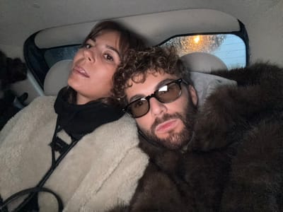 A man with curly hair and sunglasses poses next to a woman inside a car, under hanging coats. The man has a beard and a light-colored hoodie is partially visible. Rain streaks the car's rear window, reflecting blurred lights. The woman has dark hair and wears a dark top. The coats are various shades of brown, tan, and black, hung on what appears to be a coat rack.