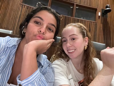 A close-up image depicts two young adult females indoors. The woman on the left has dark hair, wears a light blue and white striped button-down shirt with the collar open, and silver hoop earrings. She is making a "duck face" expression with puckered lips and her right hand rests against her cheek. The woman on the right has light brown, curly hair tied back, wears a white short-sleeved t-shirt with a graphic design partially visible, and gold-toned hoop earrings and a delicate necklace. She is smiling broadly. Her left arm is raised with the hand mostly out of frame.
The background features dark wooden walls with vertical slats. In the upper left background, two small, rectangular window-like openings or cutouts are visible. In the upper right, a dark-colored camera or recording device is mounted on the wall. Dark seating is partially visible behind the individuals. The setting appears to be an interior academic space, such as a lecture hall or seminar room, consistent with facilities found within Princeton University in Princeton, New Jersey, United States.