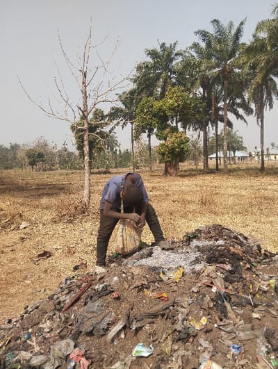The scene, located in Unguwan Rimi, Nigeria, depicts an outdoor environment under a clear sky. A young male, wearing a blue shirt and dark trousers, is positioned bent over a large refuse pile in the foreground. He is actively engaged with the pile, holding a light-colored bag, appearing to collect items from the mixed debris, which includes soil, ashes, plastics, and other discarded materials. The ground is dry and brown with sparse vegetation. A tall, leafless tree is present in the mid-ground to the left, and a line of palm trees stretches across the distant background, where several light-colored buildings with blue roofs are also visible.