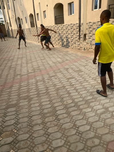 An outdoor scene in Eziobodo, Nigeria, depicts five adult males on a paved surface composed of interlocking light grey/beige hexagonal and rectangular pavers, interrupted by a horizontal stripe of red pavers. A light-colored building with multiple windows, some arched openings, and a section of dark brick-patterned wall forms the background. Four shirtless males in shorts are actively engaged in playing football (soccer) barefoot, with a visible ball centrally positioned among two of them. One male, positioned further left, observes the play, while another is partially visible on the far left. In the foreground, facing away from the camera, a fifth male stands wearing a yellow short-sleeved shirt with blue trim, black shorts with white stripes, and dark slide sandals, observing the game. Bright daylight casts distinct shadows from the individuals onto the paved ground and from the building features.