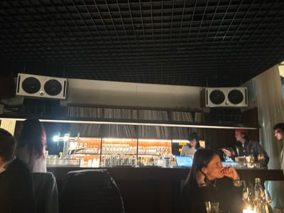 The scene depicts the interior of a dimly lit establishment, identified as a bar or restaurant located in Uccle - Ukkel, Belgium. The overhead ceiling is a dark, grid-patterned acoustic tile system. Two large, rectangular white speaker enclosures, each containing two visible circular drivers, are suspended from the ceiling, one on the left and one on the right side of the frame.
Below the speakers, a long, recessed shelf spans the background, filled horizontally with numerous vinyl records, indicating a prominent music collection. Below this shelf, a well-stocked bar counter is visible. The bar's back wall features illuminated shelving displaying various glasses and bottles. On the left side of the bar counter, a chrome beer tap system with a bright light is present.
In the foreground, a woman with dark hair, wearing a black top and silver bracelets, is seated at a dark table, facing right in profile. Her right hand is near her chin. A clear glass and a small, flickering light source (possibly a candle) are on the table in front of her. Partially visible to the far left is the back of another person's head with reddish-purple hair.
Behind the bar counter, three individuals are present. To the left, a person wearing a light-colored top and a dark baseball cap is positioned behind a laptop displaying a blue screen. In the center-right, a person with longer hair, wearing a red bandanna and a dark jacket, stands holding a small electronic device. To the far right, the head and shoulder of a man with dark, curly hair are visible as he faces left, seemingly interacting with the person in the red bandanna. The overall lighting is subdued, with accent lighting on the bar shelves and foreground table contributing to a warm ambiance.