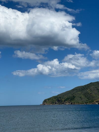 The image shows a view of M'Diq ⵎⴹⵢⴰⵇ المضيق, Morocco. The right portion of the image is dominated by the sea. Adjacent to the sea is a steep, green, mountainous coastline. The left portion of the image contains a blue sky and fluffy, white clouds.
