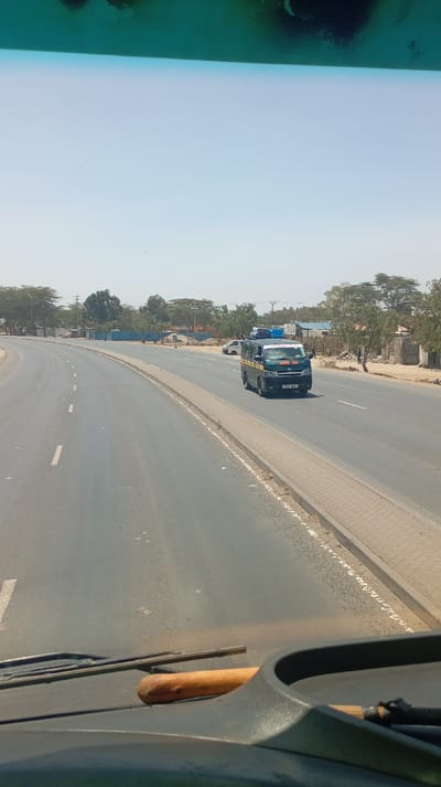 The image depicts a daytime scene from the interior of a vehicle, looking out onto a multi-lane asphalt road in Viwandani ward, Kenya. The foreground shows a dark vehicle dashboard, with windshield wipers at the base of the windshield and a light-colored wooden stick or handle resting horizontally on the dash. The top portion of the frame is partially obscured by a dark green windshield tint and smudges.
The paved road features distinct white dashed lane markings and a central median with a concrete curb and a patterned light-colored sidewalk on the right. A dark-colored passenger van, possibly a matatu, with luggage secured to its roof rack, is observed traveling away from the camera on the right side of the road. A white car is partially visible behind the van. Further in the distance, additional vehicles are present.
The environment includes clear blue skies above. Both sides of the roadway are lined with scattered trees, characterized by their light green, sparse foliage, consistent with a dry or semi-arid region. On the right side of the road, beyond the sidewalk, structures, including low-rise buildings and what appears to be a construction site with blue barriers and disturbed earth, are visible. Multiple figures, consistent with pedestrians, are discernible on the far right sidewalk/shoulder in the mid-ground.