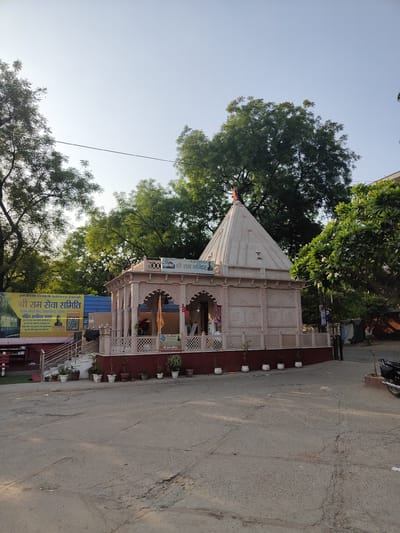 The image captures a serene view of the Shri Ram Mandir, a temple situated in Gurgaon, India. The temple, constructed from pale pink stone, features intricate carvings and a conical spire topped with an orange flag. A white railing encloses a small courtyard in front of the temple, adorned with potted plants.
To the left of the temple, a banner displays information about the Shri Ram Seva Samiti, an organization dedicated to serving the community. The banner also includes a photograph of a man and a QR code, likely for donations or further information.
In the background, lush green trees provide a natural backdrop to the temple. The sky above is a clear, light blue, indicating a pleasant day. The foreground shows a paved road with some cracks, suggesting a typical urban setting. A motorcycle is parked on the right side of the image. The overall scene exudes a sense of peace and spirituality.