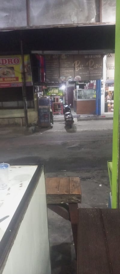 The image shows a street scene in Kecamatan Waru, Indonesia (latitude: -7.35, longitude: 112.764). The scene is viewed from a low angle, looking out from a partially open doorway. A white refrigerator-like object and a wooden bench are in the foreground. The street is paved with asphalt and there is a small, dark-colored motorcycle parked near the right side of the frame. The motorcycle is facing toward the viewer. There is a person obscured by the white refrigerator-like object, near the parked motorcycle. Behind the street is a row of buildings with signs in Indonesian. One sign reads "WARUNG Bu Rara." The buildings have windows, wooden doors, and some have awnings. The lighting is dim and there are shadows. The scene appears to be at night.