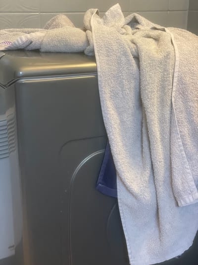 The image depicts an indoor scene in Fegersheim, France, focusing on a light grey appliance, likely a washing machine or dryer. Draped across the top and cascading down the right side of the appliance are several off-white or light cream terry cloth towels. A segment of a light purple or lavender towel is visible on the right side, tucked beneath one of the larger towels, extending down the appliance's side. The background features white rectangular tiles on a wall directly behind the appliance, and a portion of a white, vertically ribbed object is visible on the far left. The lighting is soft, highlighting the texture of the towels and the reflective surface of the appliance. No individuals are present.