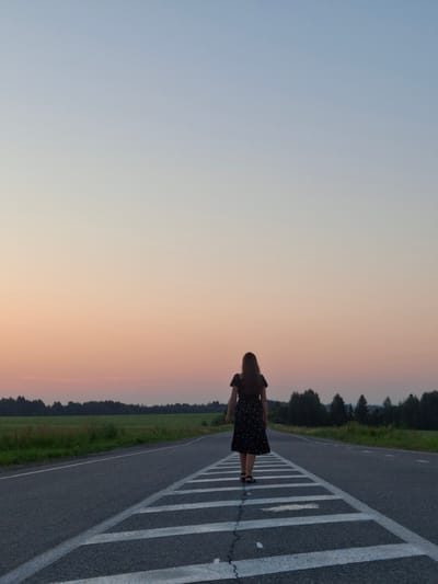 The image depicts a solitary young woman from behind, walking on an asphalt road at dusk. She is wearing a dark, short-sleeved dress with a small white pattern and dark sandals. The road has white dashed lines marking a crosswalk or pedestrian crossing. The background features a flat landscape with a line of dark green trees and a field of green grass. The sky above is a gradient of soft blue and pale orange hues, indicating the setting sun. The location is specified as Perevoznoe, Russia.