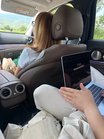 The image captures the interior of a car, viewed from the rear passenger side, looking forward. In the foreground, a person's hands and lap are visible, holding an open silver Apple MacBook laptop. The laptop screen displays the Google search page, with the "Google" logo and search bar clearly visible, appearing inverted from the camera's perspective. This person is wearing light-colored clothing, including what appears to be pleated fabric on their arm and light-colored trousers or a skirt.
In the middle ground, the back of a brown leather car seat and headrest is prominent. Seated in the front passenger seat is another individual, whose back of the head and shoulder-length light brown hair are visible. They appear to be wearing glasses and a white t-shirt, looking towards the front of the vehicle. A light-colored handbag rests on the floor or seat beside them.
Through the car windows, a bright, sunny daytime scene is visible, featuring lush green trees and distant hills, suggesting a natural and possibly rural or mountainous landscape. The car's interior details include chrome air vents in the back console, a cream-colored sun visor with an "AIRBAG" warning label and associated safety symbols, and brown leather upholstery throughout. The setting indicates a journey in a modern car, likely through a scenic area, with the provided location being Δημοτική Ενότητα Προμαχώνος, Greece.