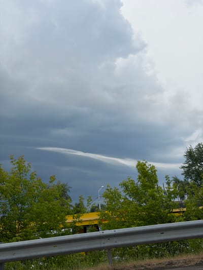 The media file shows an outdoor scene with a cloudy sky. There are trees with green leaves in the foreground and midground, some of which are deciduous. A yellow structure, possibly a building or a vehicle, is partially visible behind the trees. A metal guardrail runs along the right side of the image, separating the scene from a grassy roadside. The sky is overcast with various cloud formations, including some that appear wispy and stretched out. The lighting suggests it is daytime, but the heavy cloud cover may indicate a subdued light, possibly before or after rain. There are no visible people or text in the image. The overall mood is natural and somewhat somber due to the overcast sky. Given the location context of Tomsk, Russia, this could be a typical view of the natural landscape or roadside scenery in the region during a cloudy day.