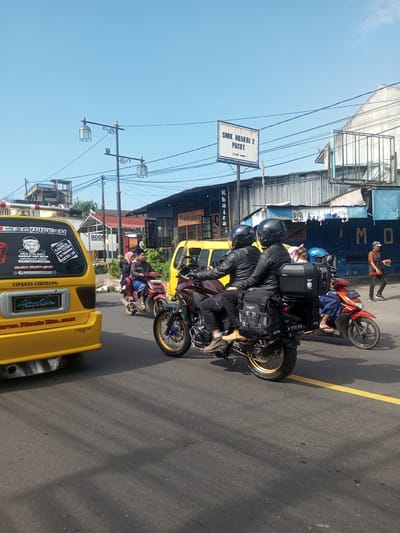 A street scene in an urban area of Indonesia on a sunny day. The view is at street level. Two people on a red and black motorcycle with black helmets and black leather jackets are in the center. The motorcycle has a luggage carrier box and side bags. Other motorcycles are nearby, with additional people on them. There's a yellow van in the foreground, which has many stickers on its rear window, and a sign on the top of the van. The street has a yellow dividing line. Behind the van and motorcycles, there are several shops and buildings, and some pedestrians on the sidewalk. A sign for "SMK NEGERI 2 PACET" is visible. Electric poles and wires are overhead.