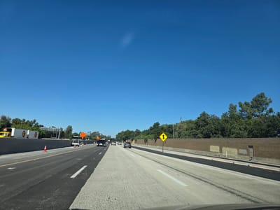 The image captures a view of a highway with ongoing construction in West Sacramento, California. The highway is divided into multiple lanes, with some sections appearing freshly paved with asphalt and others with a concrete surface. Traffic cones are visible along the sides of the road, indicating lane closures or construction zones. Several vehicles, including cars and trucks, are on the highway, navigating the construction. On the right side of the highway, a brick wall serves as a barrier, with a door set into it. Beyond the wall, lush green trees form a dense line, suggesting a natural landscape bordering the urban area. The sky above is a clear, vibrant blue, indicating a sunny day. The overall scene depicts a typical highway environment with the presence of construction and road work.