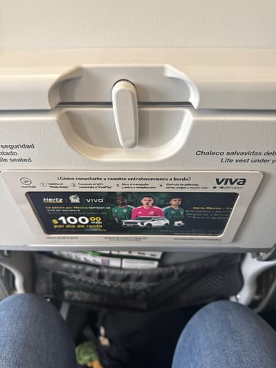 The image shows an airplane tray table in the lowered position. On the tray table is an advertisement card for Viva Aerobus, a Mexican airline. The advertisement provides instructions on how to connect to the airline's entertainment system and advertises a rental car deal with Hertz in Mexico for $100.00 MXN per day. The text on the tray table indicates the location of the life vest. A person wearing blue jeans is sitting in the adjacent seat. The location appears to be inside of an airplane. There is nothing to suggest any of this image was taken in San Antonio.