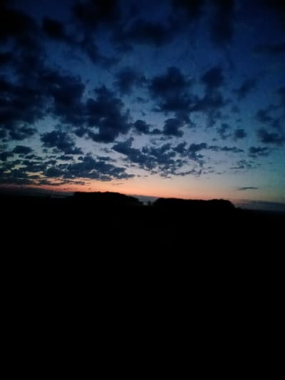 The image depicts a dusk or dawn sky over a silhouetted landscape. The sky is a deep blue, transitioning to lighter shades of blue and a band of orange and pink near the horizon. Scattered, dark clouds are visible against the sky. The foreground is entirely black, obscuring any details of the ground or any structures or natural features that might be present. There are no people, animals, or discernible objects in the frame. The scene is devoid of any activity or interaction. The location is stated to be Musakaeva, Russia, suggesting an outdoor, possibly rural or open area setting.