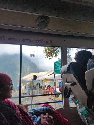 The image shows the interior of a bus, likely in Burwa, India, looking out a window at a cloudy, mountainous landscape. A woman wearing glasses and a red headscarf is seated on the left, looking out the window. A child is sleeping on her lap, covered by a blue and pink blanket. To the right, another passenger's head is visible, facing away from the camera. Outside the window, there are tents, stalls, and chairs, suggesting a roadside market or rest stop. A green sign with white text is visible, reading "BORDER ROADS ORGANISAT WE ARE ON DUT SO THAT, YOU ENJOY THE BE." A plastic water bottle is on the seat to the right. The bus ceiling has a circular speaker. The overall scene depicts travel in a mountainous region.